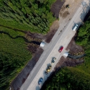 An aerial view of newly replaced culvert on a dirt road in rural Alaska. Cars and construction machines are parked on the road above the culvert, and a stream of water cuts through the greenery of pine trees and foliage.