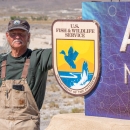A man with a white mustache wearing overalls and a U.S. Fish and Wildlife Service volunteer cap standing next to a sign with the U.S. Fish and Wildlife Service logo on it