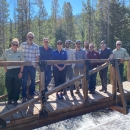 group of 10 people standing on a bridge above a fast flowing creek in the woods
