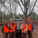 Four people in orange hunting vests, two with firearms in their hands, stand in front of a truck in the woods smiling.