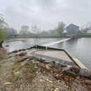 A metal fish ladder on a concrete slope next to a low-head dam near the Stockdale Mill on a cloudy day.