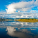 Still water providing a mirror image of the clouds and blue sky above with vegetation on both left and right banks of the water with mountains on the horizon. 