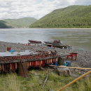 Fish camp along the banks of a river with forest covered hills in the background and large drying racks on the shoreline filled with processed salmon. 