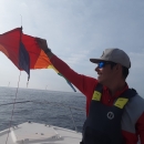 A person wearing a red shirt, black flotation vest and tan cap holds a yellow, red and orange kite aloft while standing in the bow of a boat