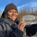 A woman wearing a beanie and jacket holds up a clear container with water and a small aquatic organism