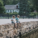 Two young boys sit on the edge of a stone walled pond and feed the trout and ducks. The historic 1899 hatchery building is visible in the background.
