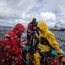 People in bright orange and yellow rain gear in an orange skiff boat through choppy waters through a rain spattered lens. A blue and white ship is in the background.