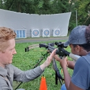 A man teaching a woman how to handle a crossbow while she aims at an archery target