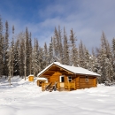 A wood cabin stands in the snow at the edge of a forest.