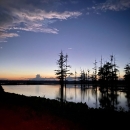 A thin orange band glows above a flat body of water, with some tall trees in the background.