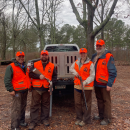 Four people in orange hunting vests, two with firearms in their hands, stand in front of a truck in the woods smiling