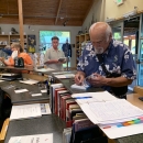 A man with white hair and beard and wearing a Hawai'ian shirt signs a book at the counter of a visitor center store.