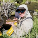 A person wearing outdoor field gear sits beside a grassy streambank, holding an Apache trout over a yellow measuring board. The stream and dense vegetation fill the background.