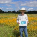 A person wearing a wide‑brimmed hat stands in a large field filled with yellow wildflowers. They hold a sign that reads “Cooperator – Partners for Fish & Wildlife – U.S. Fish & Wildlife Service.” Trees and a blue sky with scattered clouds appear in the background.
