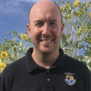 Color photo of Daniel Elbert wearing a black polo shirt with US Fish and Wildlife Service emblem on the chest. Daniel has sunflowers in the background and is smiling.