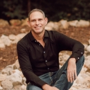 Photo of Matt with dark top and blue jeans smiling in a seated pose.