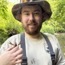 Levi in river with sun hat on and tan work shirt holding crayfish