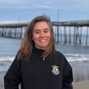 Angelina Yost smiles at the camera wearing a UFWS jacket, standing on a beach in front of a wooden pier
