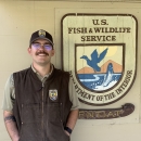 Man with mustache wearing short-sleeved Service uniform with vest and ballcap stands beside a USFWS shield sign