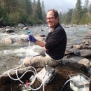 A smiling man in glasses holds scientific equipment while sitting on rocks by a stream.