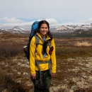A woman wearing a large backpack stands before tundra and low-lying snow-speckled mountains.
