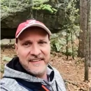 Blane smiles in a red baseball cap and grey hoodie in a forest