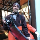 A man smiles big while holding a very big fish. He wears orange waders and a brown jacket and beanie.