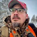 A portrait of Bryson Jones in a snowy landscape outfitted with binoculars and other hunting gear