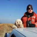 a man stands in the back of a truck with a fluffy white dog wearing a hat and orange jacket