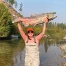a woman in a long sleeve orange shirt and tan waders raises a large spawned out salmon above her head