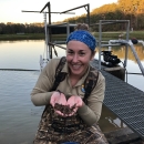 Woman in brown camo waders in a pond holds juvenile mussels cupped in her hands.