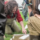 Female biologist kneels on the ground among children, while pointing to insects in a white tray