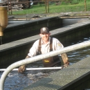 a man wearing a hat and waders stands waist deep in water while smiling at the camera.