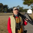 Man looking at the camera wearing a bright red long-sleeved shirt and a cream vest. He is holding a tripod over his shoulder and wearing binoculars strapped around his neck.