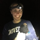 a woman wearing a headlamp in the dark holds a bat in gloved hands