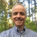 Keith smiles in a blue plaid button down in a forested area