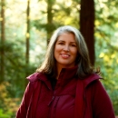 Photo of woman in a burgundy colored coat smiling, posing for photo in front of fir trees with sun shining through the trees 