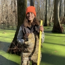 FWS staff member in cypress swamp holding measuring stick and blaze orange hat
