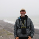 Man standing along Alaska coastline with binoculars around neck