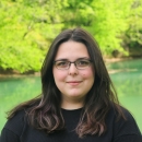 Maria smiles with glasses in a black blouse in front of a body of water and green trees