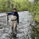 A person wearing wader overalls stands mid-calf in a river. They hold a fish in their hand with the other at their side while they smile.