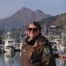 woman in USFWS uniform and sunglasses in front of a harbor with mountains in the background