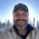 man in ballcap smiles at the camera in a spruce forest