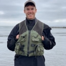 Fish Biologist, Nate Queisser, standing wearing black waterproof pants and jacket with a green multipocket vest and a tan baseball cap. Nate is standing in front of a body of water.