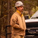 A man wearing a yellow fire uniform and hardhat stands next to a truck