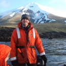 Man weariing orange float coat stands in skiff with snow-covered volcano in the background.