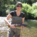 Brook Silver, Fish Biologist, wearing tan waders, black t-shirt and holding a salmonid. She is standing in a shallow creek. 
