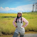 A woman wearing waders and a hat stands with her hands on her hips in a salt marsh