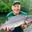 Todd smiling and holding a fish in hand infront of a river and green trees 