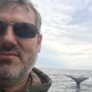 a selfie of a white man in a boat with a whale's tail emerging from the water behind him
